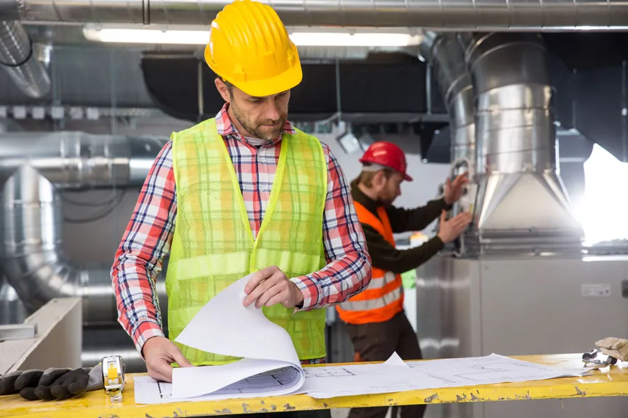 Engineer in a hard hat and safety vest reviewing plans with a colleague in a mechanical room